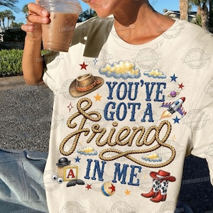 May include: A cream-colored t-shirt featuring the blue text "YOU'VE GOT A FRIEND IN ME" surrounded by a brown rope graphic, a cowboy hat, a rocket ship, a toy block with the letter A, a ball, and red cowboy boots with white stars.
