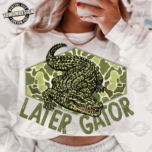 May include: White sweatshirt featuring a green alligator illustration with the text "LATER GATOR". The alligator is set against a green camouflage background within a hexagon shape. The design is a digital file.