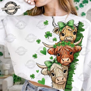 May include: White sweatshirt featuring a graphic of three Highland cows with green bows and shamrocks. The cows have different shades of brown and cream fur. The text "TUMOMDESIGN" is printed on the top left.