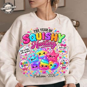 Puede incluir: Una sudadera color crema con un gráfico colorido que incluye el texto "THE YEAR OF SQUISHY HUNTING" y "Madison - 2026". El diseño presenta varias formas de dibujos animados sonrientes de colores arcoíris como estrellas, corazones y cubos.
