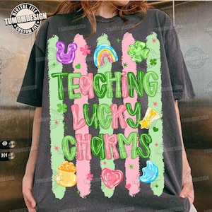 Puede incluir: Camiseta gris oscuro con las palabras "TEACHING LUCKY CHARMS" en verde y rosa, decorada con gráficos del Día de San Patricio. El diseño incluye un arcoíris, tréboles y una olla de oro.