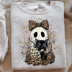 May include: A light beige sweatshirt featuring a Halloween-themed graphic. The design includes a ghost wearing a leopard print bow and holding a black cat. Bats, spiderwebs, and stars complete the spooky design.