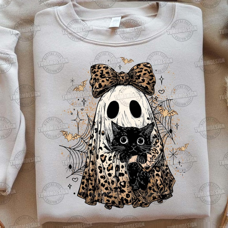 May include: A light beige sweatshirt featuring a Halloween-themed graphic. The design includes a ghost wearing a leopard print bow and holding a black cat. Bats, spiderwebs, and stars complete the spooky design.