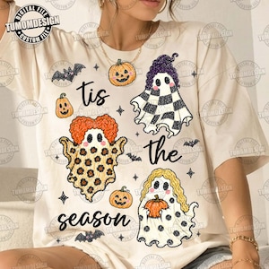 May include: A cream-colored t-shirt featuring a graphic print with the text 'tis the season'. The design includes three ghosts with different hair styles and patterns, small orange pumpkins, and black bats scattered across the front.