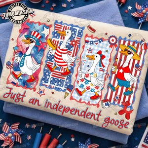 Può includere: Un asciugamano beige con quattro oche dei cartoni animati vestite con abiti patriottici. Ogni oca indossa occhiali da sole e tiene in mano oggetti come bandiere e hot dog. Il testo "Just an independent goose" è scritto in basso.