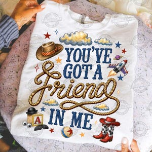 May include: A white t-shirt featuring the text "YOU'VE GOT A FRIEND IN ME" in blue letters. The design includes a brown cowboy hat, a coiled rope, a wooden block with the letter A, a ball, red cowboy boots, clouds, stars, and a rocket ship.