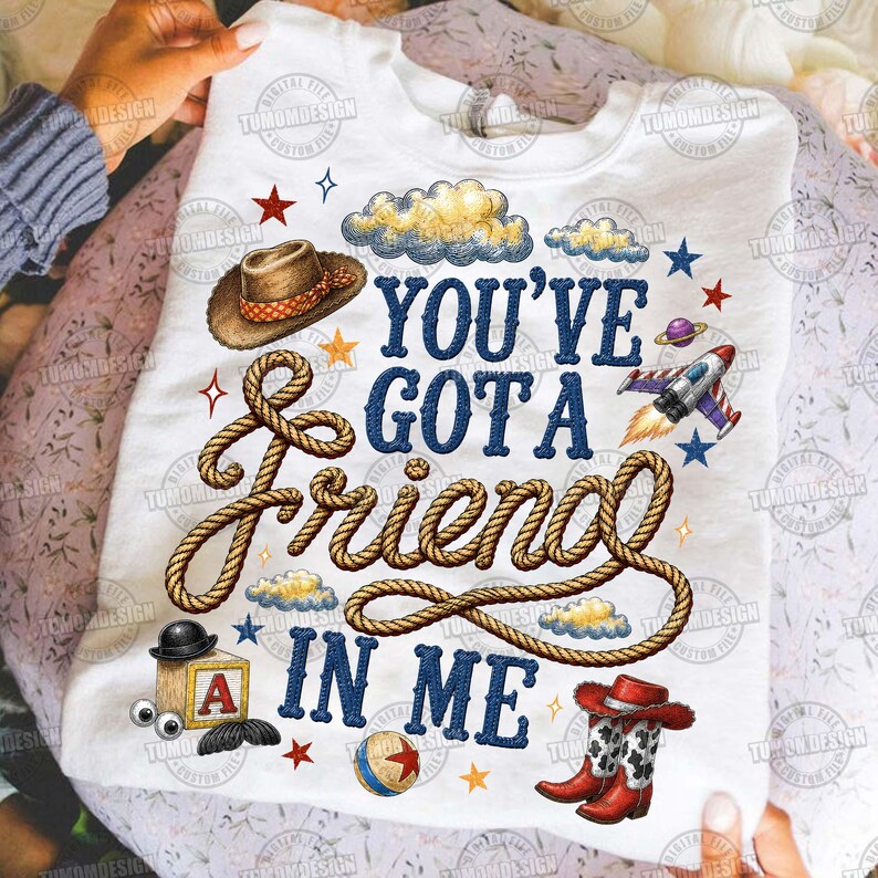 May include: A white t-shirt featuring the text "YOU'VE GOT A FRIEND IN ME" in blue letters. The design includes a brown cowboy hat, a coiled rope, a wooden block with the letter A, a ball, red cowboy boots, clouds, stars, and a rocket ship.