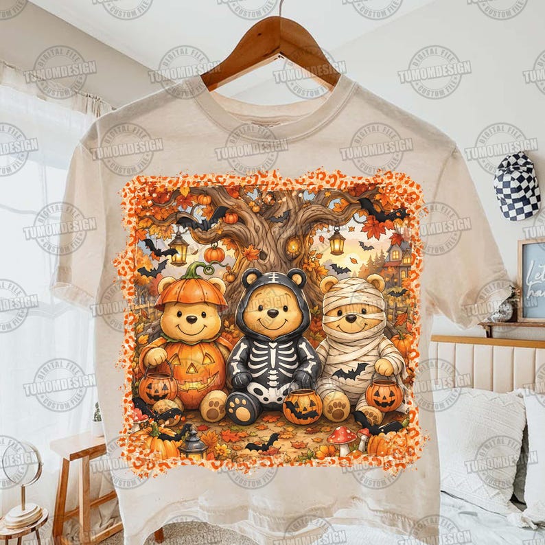May include: A beige t-shirt featuring a graphic print of three cartoon bears dressed in Halloween costumes. One bear wears a pumpkin suit, one a skeleton outfit, and one is wrapped like a mummy. The scene includes a tree, bats, and pumpkins.