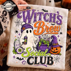 May include: A white sweatshirt featuring a graphic print with the text Witch's Brew Social Club. The design includes a ghost wearing a purple beanie, a tombstone, a green eyeball, a purple cauldron with green liquid, and a jack-o'-lantern.