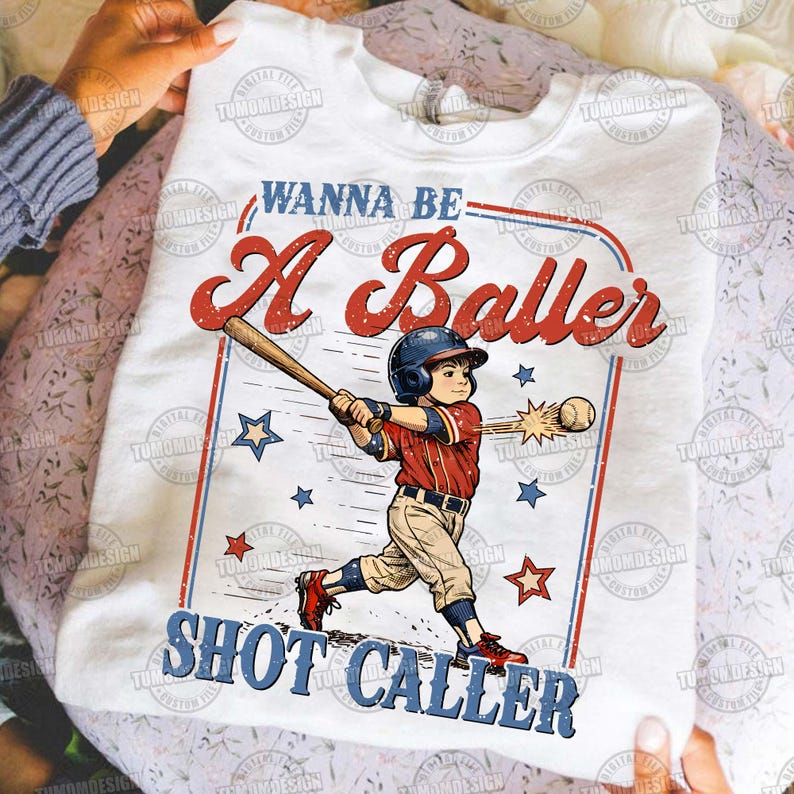May include: White t-shirt featuring a baseball-themed graphic. The design includes a baseball player swinging a bat, stars, and the text "Wanna Be A Baller Shot Caller" in red and blue. The shirt is held up by a person.