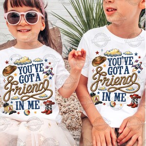 May include: Two white t-shirts featuring a graphic print with the text "YOU'VE GOT A FRIEND IN ME" in dark blue lettering. The design includes a brown cowboy hat, a coiled rope, a rocket ship, a toy block with the letter A, and cowboy boots.