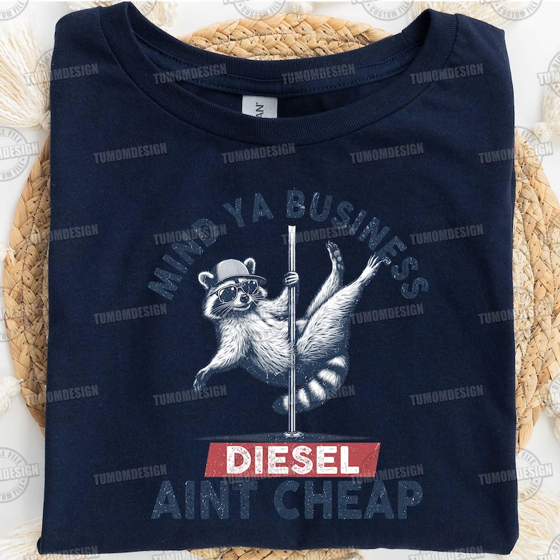May include: A dark blue t-shirt with a raccoon wearing sunglasses and a hat, holding a pole. The text "Mind Ya Business Diesel Ain't Cheap" is printed on the shirt.