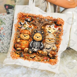 May include: A white canvas tote bag featuring a colorful Halloween-themed illustration of three bears dressed in costumes. One bear wears a pumpkin suit, one a skeleton outfit, and one is wrapped like a mummy, all surrounded by autumn leaves and bats.
