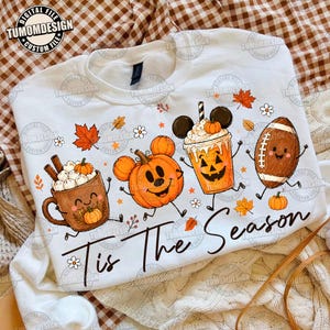 May include: A white crewneck sweatshirt featuring a graphic print of a smiling mug of hot cocoa, a pumpkin, a jack-o'-lantern drink, and a football, all with arms and legs. The text 'Tis The Season' is printed in black script below the illustrations.