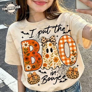 以下が含まれることがあります： I put the BOO in Boujeeというテキストがプリントされたクリーム色のTシャツ。水玉模様の蝶ネクタイをしたお化け、花柄やチェック柄のオレンジ色の文字、ヒョウ柄やチェック柄の2つのカボチャ、クモの巣、コウモリがデザインされています。