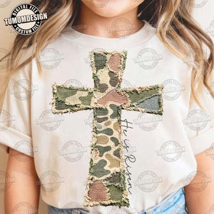 Puede incluir: Camiseta color crema con un diseño de cruz grande hecho de parches de tela de camuflaje. La cruz tiene un borde deshilachado y las palabras "He is Risen" están escritas en escritura negra debajo. La camiseta tiene mangas cortas.