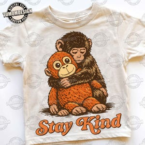 May include: Cream-colored t-shirt featuring a graphic of two cartoon monkeys hugging. The orange monkey is being embraced by a brown monkey. The text "Stay Kind" is written in orange script below the monkeys. The shirt has the text "TUMOMDESIGN" printed on it.