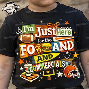 May include: Black t-shirt with a colourful graphic that reads "I'm Just Here for the Food and Commercials." The design includes a football, pizza, burger, and a football helmet. The shirt is for football fans.