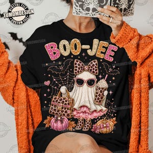 May include: A black t-shirt featuring a graphic of a ghost wearing a leopard print bow, sunglasses, and a pink skirt. The ghost holds a leopard print handbag and a drink. The text BOO-JEE is printed above, surrounded by spiderwebs and pumpkins.