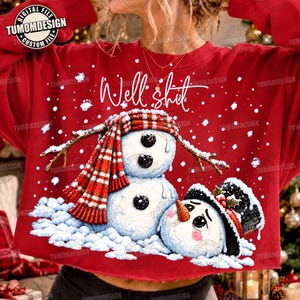 May include: A red sweatshirt featuring a graphic of two snowmen, one standing and one fallen, with the text "Well shit" in white script. The standing snowman wears a red and white plaid scarf, while the fallen one wears a black top hat.