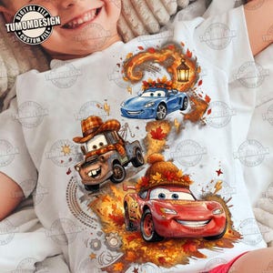 Puede incluir: Una camiseta blanca con un estampado gráfico de tres coches animados de la película Cars. El diseño incluye una grúa marrón con un sombrero de cuadros, un coche deportivo azul y un coche de carreras rojo con un gorro de punto, rodeados de hojas de otoño.