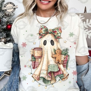 May include: A cream-colored t-shirt featuring a whimsical ghost design with a floral bow, holding a mug and gingerbread man. The ghost is draped in patchwork fabric with green and red accents, and is surrounded by Christmas lights and snowflakes.