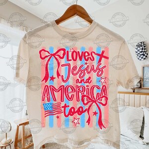 May include: Cream-colored t-shirt with the text "Loves Jesus and America too" in red script. The design includes red and blue stripes, stars, and a small American flag. The shirt is hanging on a wooden hanger.