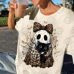 May include: Cream-colored t-shirt featuring a Halloween-themed graphic. The design includes a ghost wearing a leopard print bow and holding a black cat. Bats, spiderwebs, and stars complete the spooky illustration.