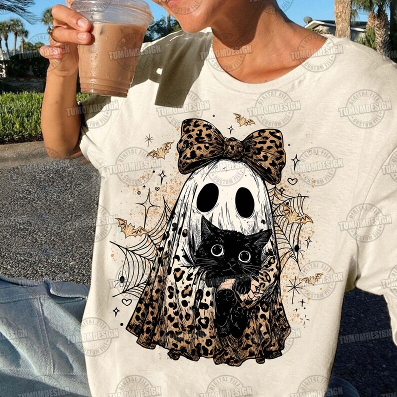 May include: Cream-colored t-shirt featuring a Halloween-themed graphic. The design includes a ghost wearing a leopard print bow and holding a black cat. Bats, spiderwebs, and stars complete the spooky illustration.
