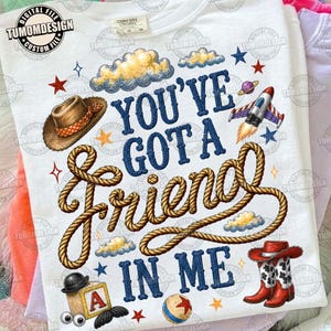May include: A white t-shirt featuring the text "YOU'VE GOT A FRIEND IN ME" in blue letters, with a rope graphic intertwined through the words. Illustrations include a brown cowboy hat, a rocket ship, a wooden block with an "A", and cowboy boots.