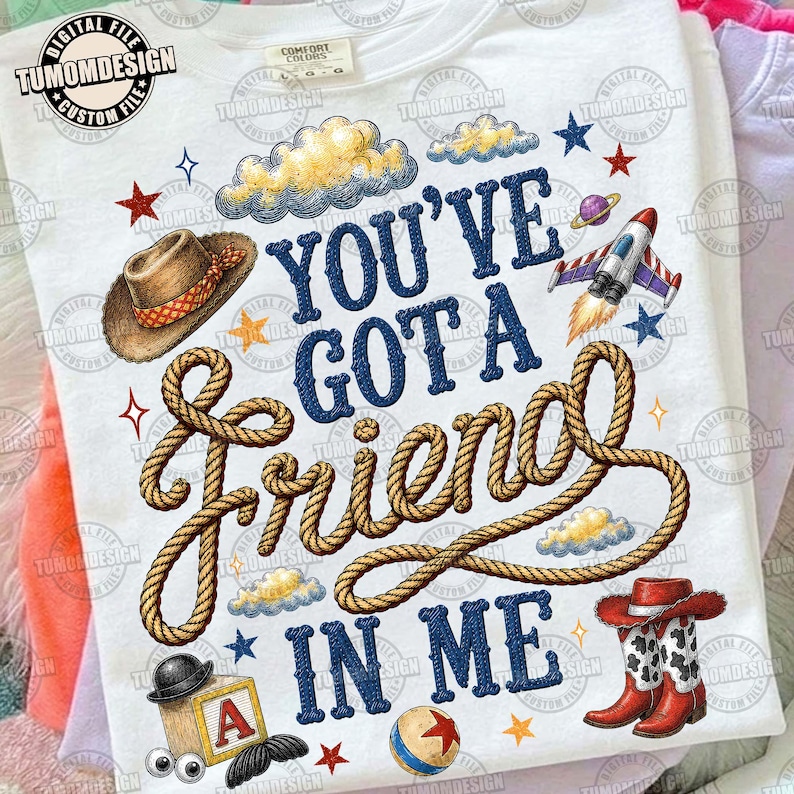 May include: A white t-shirt featuring the text "YOU'VE GOT A FRIEND IN ME" in blue letters, with a rope graphic intertwined through the words. Illustrations include a brown cowboy hat, a rocket ship, a wooden block with an "A", and cowboy boots.
