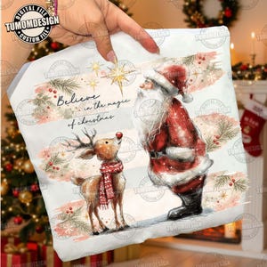 Op de afbeelding: Een wit stoffen vierkant met een aquarelillustratie van de Kerstman en een rendier met een rode sjaal. De tekst luidt Believe in the magic of christmas. Het ontwerp bevat dennentakken, rode bessen en een gouden ster op een lichte achtergrond.