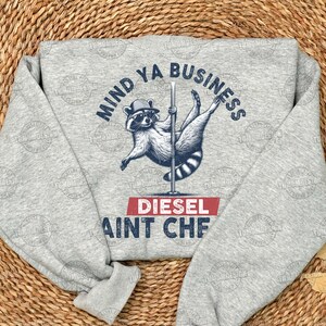 May include: Gray sweatshirt with a raccoon wearing a hat and holding a pole. The text on the sweatshirt says "Mind Ya Business Diesel Ain't Cheap".