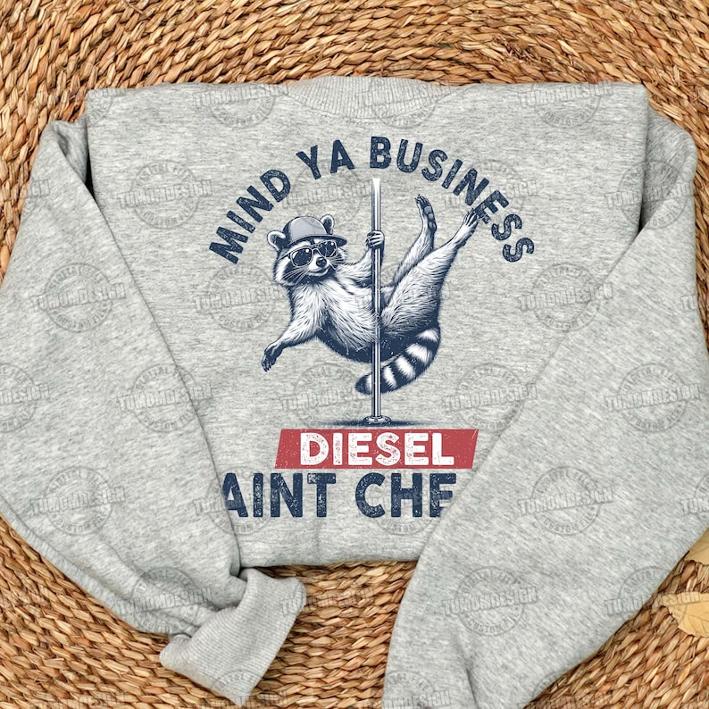 May include: Gray sweatshirt with a raccoon wearing a hat and holding a pole. The text on the sweatshirt says "Mind Ya Business Diesel Ain't Cheap".