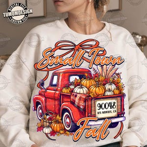 May include: A cream-coloured sweatshirt featuring a graphic of a red pickup truck filled with pumpkins and autumn foliage. The text reads Small Town and Fall, with a licence plate displaying 90048 Los Angeles, CA. The design includes a large orange bow.