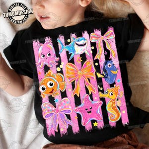 May include: Black t-shirt featuring colorful cartoon sea creatures and bows on pink striped background. Includes an octopus, shark, clownfish, bow, starfish, seahorse, and Dory. The text "TUMOMDESIGN" is visible.
