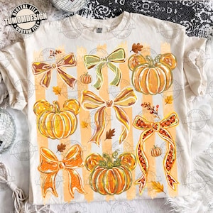 Puede incluir: Una camiseta de color crema con un patrón repetitivo de calabazas naranjas con forma de orejas de ratón y varios lazos decorativos en tonos naranja, marrón y verde. El diseño incluye pequeñas hojas de otoño y rayas verticales de fondo.