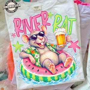 May include: A white t-shirt featuring a graphic of a rat wearing sunglasses and a floral shirt, floating in a watermelon-patterned inner tube while holding a glass of beer. The text RIVER RAT is printed in pink and green above the rat.