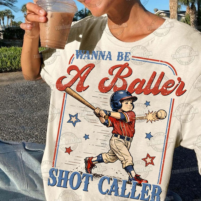 May include: Cream-colored t-shirt featuring a baseball-themed graphic. The design includes a baseball player swinging a bat, stars, and the text "WANNA BE A Baller SHOT CALLER" in red and blue. The shirt is a casual wear item.