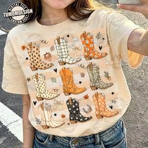 以下が含まれることがあります： クリーム色のTシャツで、ヒョウ柄、チェック柄、花柄、水玉模様、ストライプなど、様々なパターンの9足のウエスタンブーツのグラフィックプリントが施されており、葉、リボン、ハート、星、サボテンの小さなアイコンが散りばめられています。