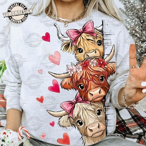 Può includere: Una felpa grigio chiaro con un design stravagante di tre mucche delle Highlands impilate verticalmente. Ogni mucca ha un aspetto unico, con fiocchi rosa, occhiali a forma di cuore e corone floreali. Cuori rossi sono sparsi. Il testo "TUMOMDESIGN" è visibile.