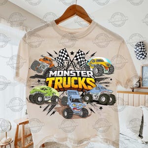 K&ouml;nnte beinhalten: Beiges T-Shirt mit einem farbenfrohen Monster-Truck-Motiv. Das Design umfasst verschiedene Monster-Truck-Illustrationen, karierte Flaggen und den Text "MONSTER TRUCKS" in einer kr&auml;ftigen, gelben Schrift. Das Shirt h&auml;ngt an einem Holzb&uuml;gel.