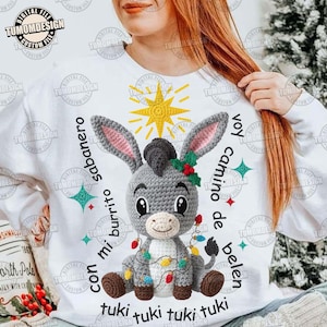 Könnte beinhalten: Weißes Sweatshirt mit einem gehäkelten Esel, einem Stern und Text. Der Esel ist grau mit rosa Ohren, braunen Hufen und einer Lichterkette. Der Text enthält "con mi burrito sabanero" und "voy camino de belen".