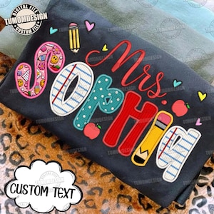 May include: A dark gray t-shirt featuring the text Mrs. Sophia in colorful, patterned letters. The design includes school-themed graphics like pencils, lined paper, apples, and smiley faces, accented with small hearts on a dark fabric background.