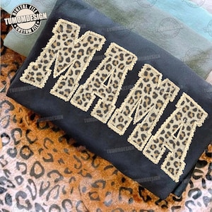 May include: Black sweatshirt with the word "MAMA" in leopard print letters. The letters have a gold-colored border. The background includes a leopard print pattern and the text "TUMOMDESIGN" in a circle.