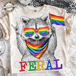 May include: Cream-colored t-shirt featuring a raccoon wearing rainbow heart-shaped sunglasses and a rainbow bandana. The raccoon is holding a rainbow flag and making a rock-and-roll hand gesture. The word "FERAL" is printed in colorful letters.