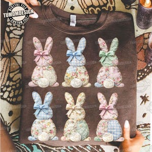 May include: A brown sweatshirt featuring six appliqued Easter bunnies. Each bunny has a different floral or plaid pattern, a bow, and a white egg-shaped tail. The text "TUMOMDESIGN" is visible in the image.