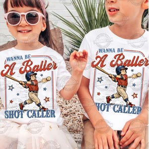 May include: Two white t-shirts with a baseball theme. Each shirt features a baseball player graphic with the text "WANNA BE A Baller" and "SHOT CALLER" in red and blue. The shirts are worn by children.