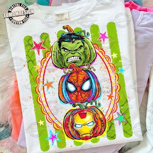May include: A white t-shirt featuring a graphic print of three stacked pumpkins designed to resemble superhero characters. The top pumpkin is green, the middle is red and blue with a web pattern, and the bottom is red and gold with a face mask.