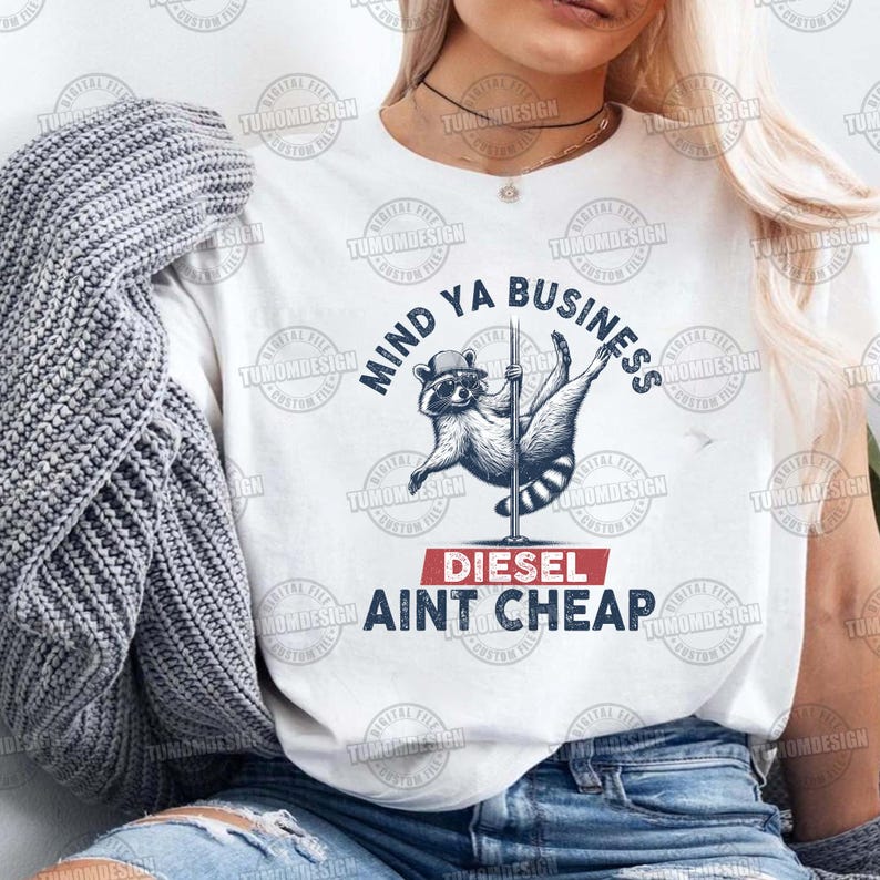 May include: White t-shirt with a raccoon wearing a hat and holding a pole. The text on the shirt reads "Mind Ya Business Diesel Ain't Cheap".
